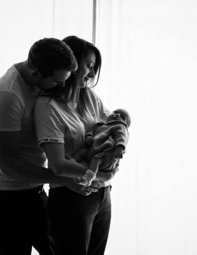 famille parents avec un nouveau né dans les bras devant une fenêtre
